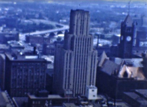 Aerial view of a city skyline featuring a tall building and clock tower, with a highway in the background.