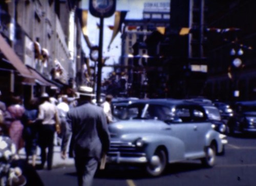 A busy street scene with pedestrians, vintage cars, and festive decorations in a city setting.