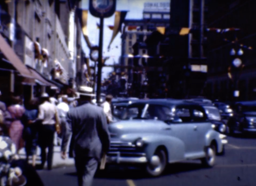 A busy street scene from the past, featuring pedestrians, vintage cars, and festive decorations.