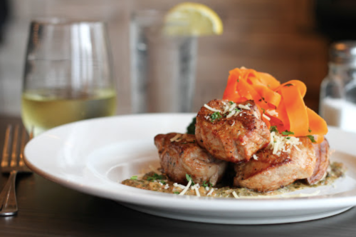 A plate of seared meat topped with carrot ribbons and herbs, served with a sauce, alongside a glass of white wine.