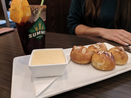 A plate with four soft pretzel bites topped with salt, served with a creamy dip, alongside a drink with an orange slice.