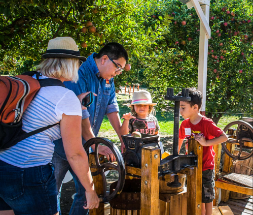 A family gathers around a wooden apple press in an orchard, with children observing and adults assisting.