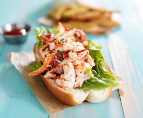 A lobster roll with lettuce on a bun, served with fries and a small bowl of sauce in the background.