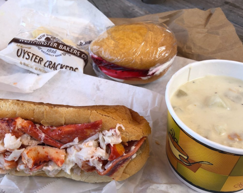 A lobster roll with a side of oyster crackers, a dessert sandwich, and a cup of clam chowder on a table.