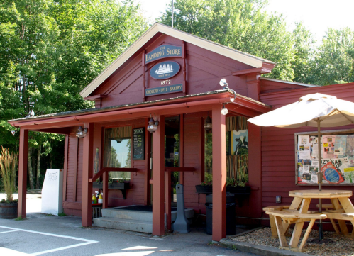A red wooden building with a sign reading "The Landing Store," featuring outdoor seating and surrounded by trees.