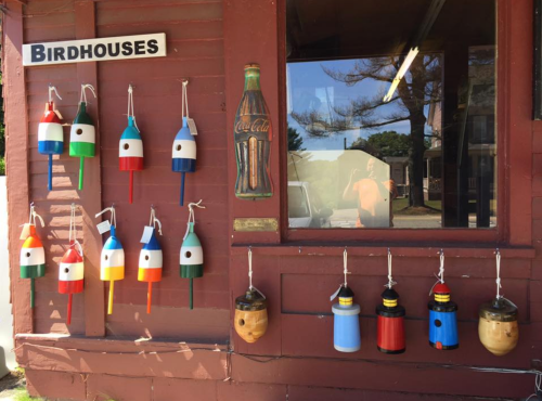 Colorful birdhouses and decorative items hanging on a wooden wall, with a vintage Coca-Cola sign nearby.