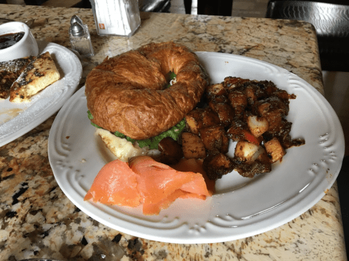 A croissant sandwich with lettuce and smoked salmon, served with seasoned roasted potatoes and a side of fruit.