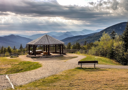 Scenic view of a gazebo on a hilltop, surrounded by mountains and trees under a cloudy sky.