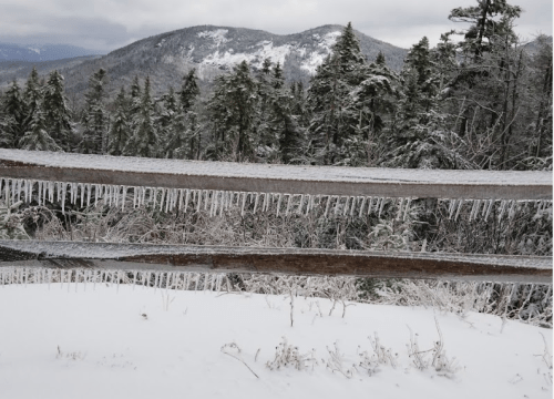A snowy landscape with a wooden fence, icicles hanging down, and mountains in the background under a cloudy sky.