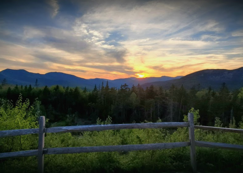 Sunset over mountains, with a wooden fence in the foreground and lush greenery below a colorful sky.
