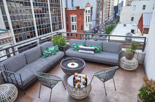 A stylish rooftop patio with striped sofas, a fire pit, and drinks, surrounded by city buildings.