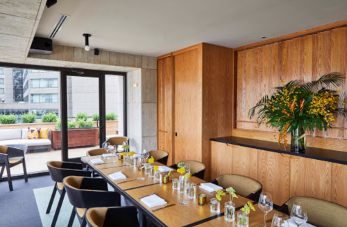 A modern restaurant interior with a long table set for dining, featuring wooden accents and vibrant floral arrangements.