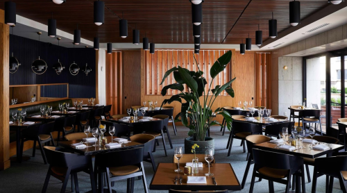 Modern restaurant interior with wooden accents, elegant tables set for dining, and a large potted plant in the center.
