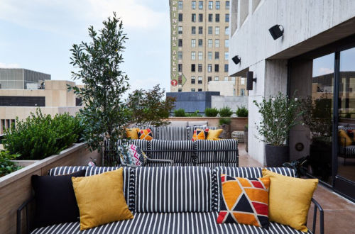 Rooftop terrace with striped sofas, colorful cushions, and greenery, overlooking a cityscape with a sign in the background.