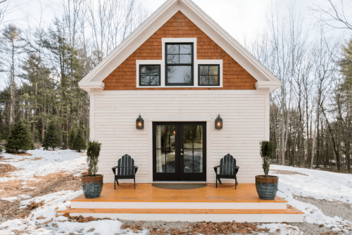 A cozy, modern cabin with a wooden porch, surrounded by snow and trees, featuring two chairs and potted plants.