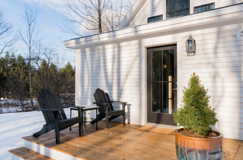 A cozy porch with two black chairs, a potted evergreen, and a door, surrounded by snow and trees.