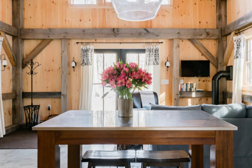 A cozy wooden interior with a dining table, floral centerpiece, and large windows letting in natural light.