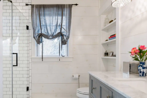 Bright bathroom featuring a glass shower, white shiplap walls, a window with gray curtains, and a stylish vanity.