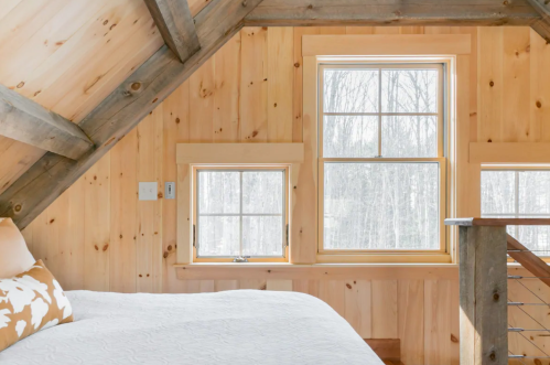 Cozy bedroom with wooden walls, two windows, and a sloped ceiling, overlooking a snowy landscape.