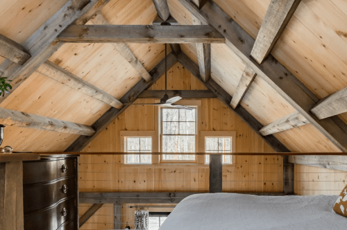 Cozy loft interior with wooden beams, large windows, and a view of a rustic bedroom below.