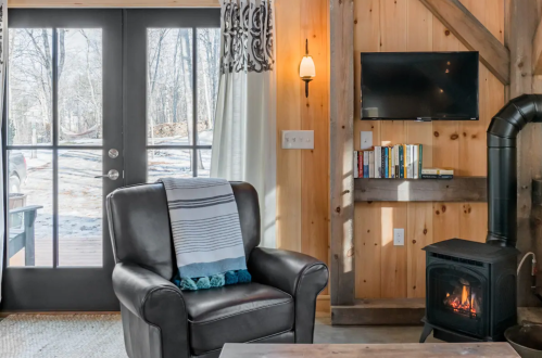 Cozy living space with a leather armchair, wood stove, and large windows showing a snowy outdoor scene.