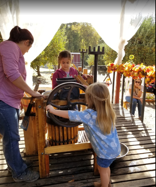 Two children and an adult operate a vintage apple press outdoors, surrounded by autumn decorations and trees.