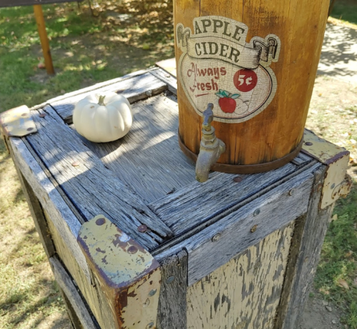 A vintage apple cider dispenser on a wooden crate, with a small white pumpkin beside it.