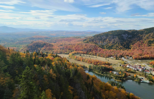 A panoramic view of vibrant autumn foliage covering hills and a river, with a clear blue sky above.
