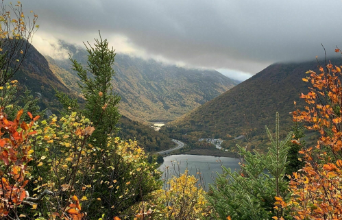A scenic view of a valley with autumn foliage, mountains, and a winding river under a cloudy sky.