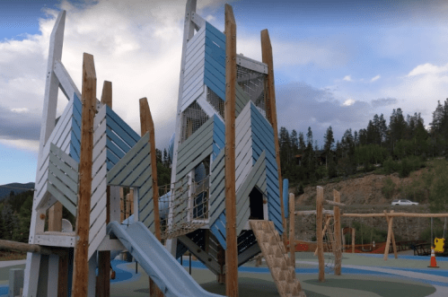 Colorful playground structure with slides and climbing features, surrounded by trees and a cloudy sky.