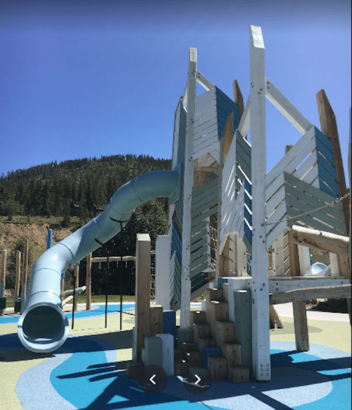 A playground structure featuring a large slide, climbing elements, and colorful ground surfaces, set against a mountain backdrop.