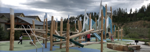 A playground with wooden structures and climbing features, surrounded by trees and cloudy skies.