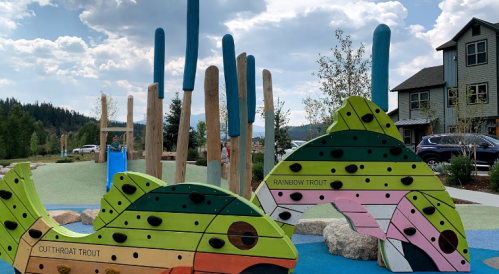 Colorful wooden fish sculptures in a playground, with trees and a house in the background under a cloudy sky.