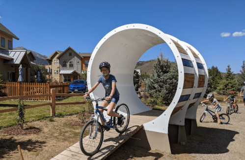 A child rides a bike through a large, white circular structure in a scenic neighborhood with houses and trees.
