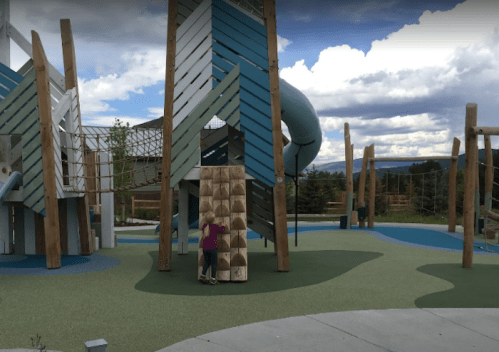 A child climbs a wooden wall at a colorful playground with slides and climbing structures under a cloudy sky.