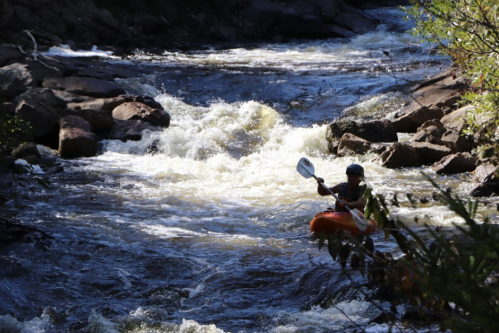 A person kayaking on a turbulent river surrounded by rocks and greenery.