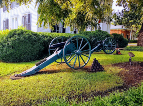 A historic cannon on display in a grassy area, with a stack of cannonballs nearby and a house in the background.