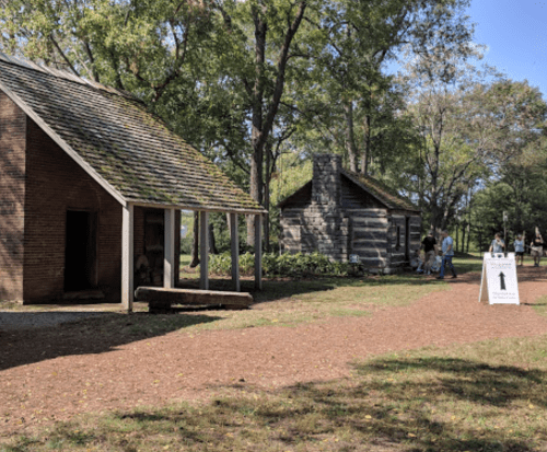 Two historic buildings in a park setting, with visitors walking along a gravel path and a directional sign nearby.