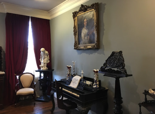 A vintage room featuring a piano, ornate decor, a statue, and a large framed portrait on the wall.