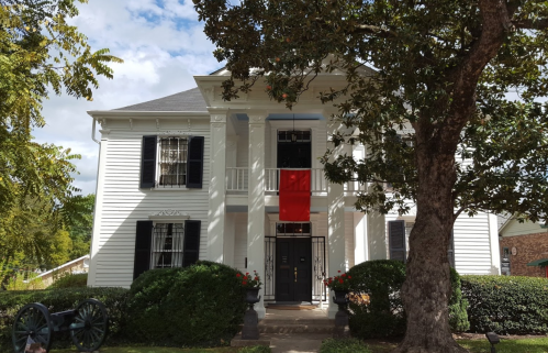 A white two-story house with black shutters, a red banner hanging from the second floor, and a large tree in front.