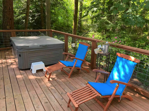 A wooden deck surrounded by trees, featuring a hot tub and two blue lounge chairs.