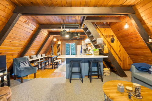 Cozy attic living space with wooden beams, a kitchen island, seating area, and stairs leading to an upper level.