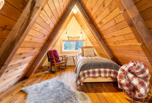 Cozy attic bedroom with wooden walls, a bed, rocking chair, and soft textiles, illuminated by warm lighting.