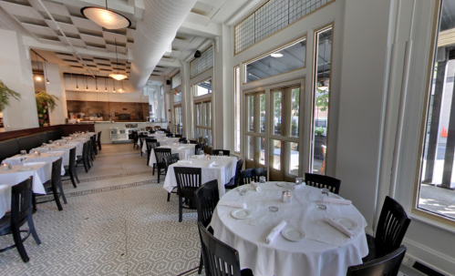 Interior of a modern restaurant featuring white tablecloths, black chairs, and large windows for natural light.
