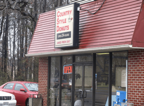 Sign for "Country Style Donuts," open 24 hours, with a red car parked outside and trees in the background.