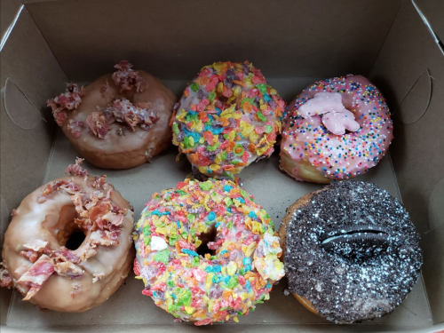 A box of six colorful donuts, including glazed, fruity cereal, and chocolate varieties, some topped with bacon and sprinkles.