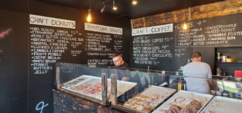 A cozy donut shop with a menu board, showcasing various craft donuts and coffee options, and two staff members behind the counter.