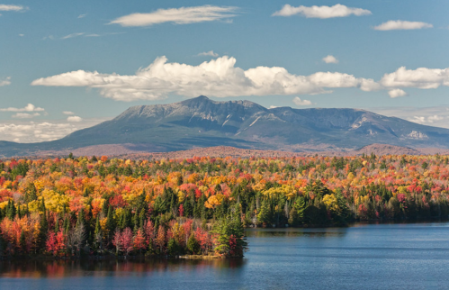 Vibrant autumn foliage surrounds a serene lake, with a majestic mountain backdrop under a blue sky with fluffy clouds.
