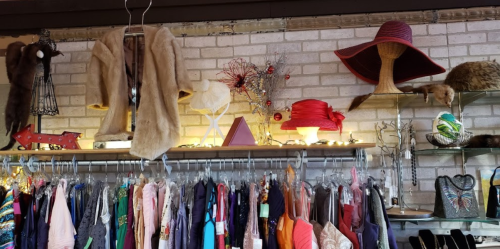A vintage clothing store display featuring dresses, hats, and decorative items on shelves against a brick wall.
