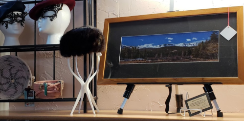 A framed landscape photo on a display shelf with a fur hat, decorative items, and mannequin heads in the background.